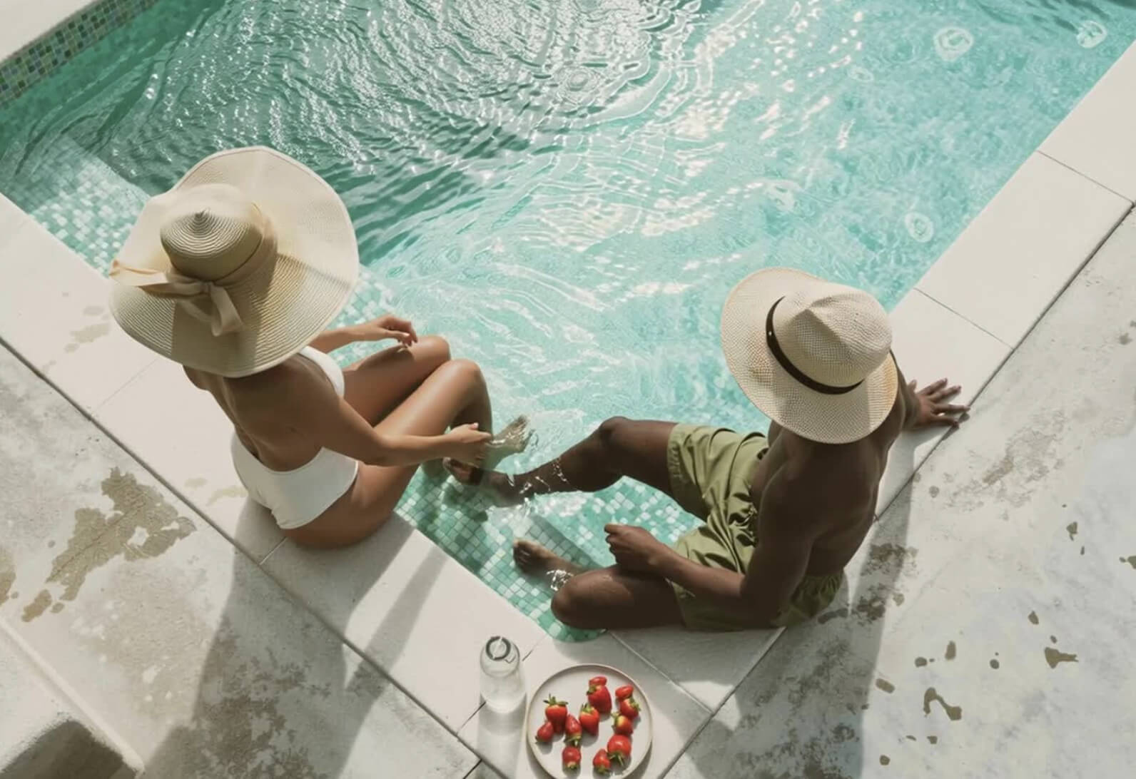 Couple sitting at the edge of The Code Austin courtyard pool wearing sun hats and sharing strawberries and water in the afternoon light.
