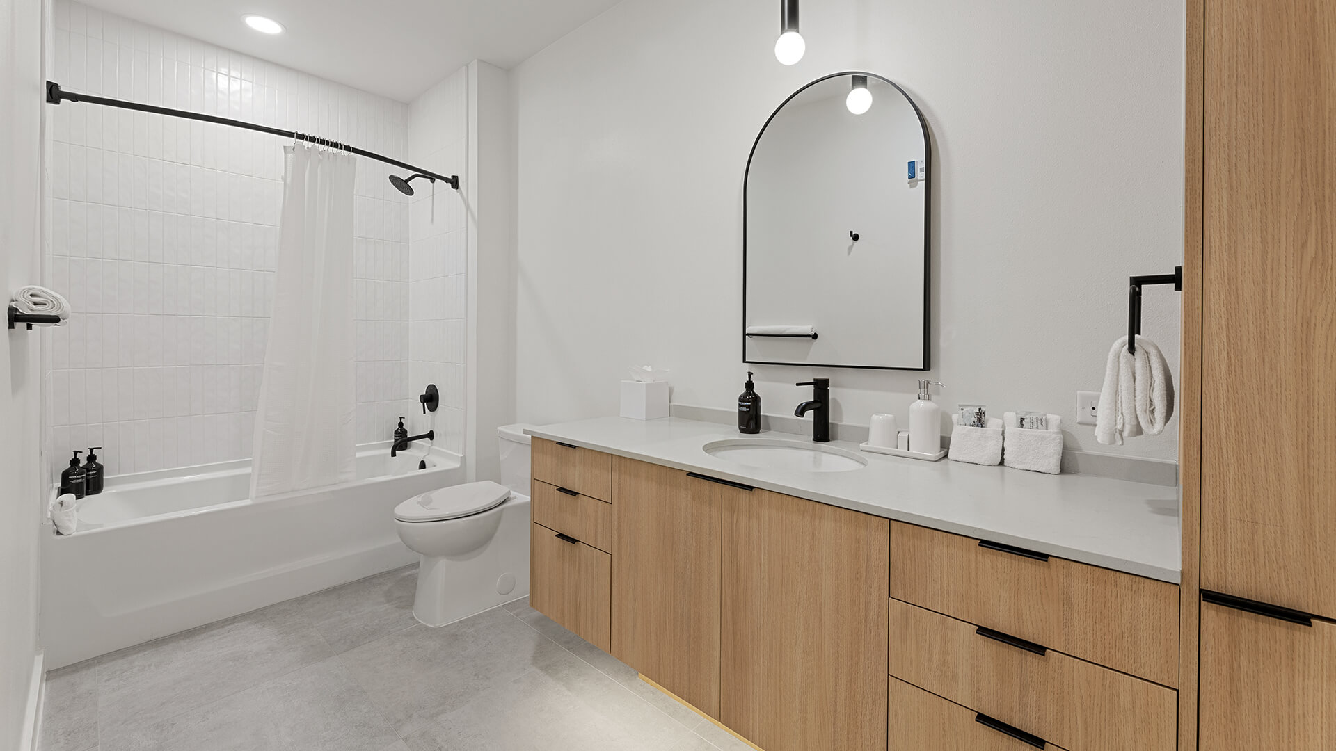 Bathroom at The Code Austin in the Shady Grove palette with medium wood cabinetry, double sink vanity, large shower with light tile, and light flooring and walls.